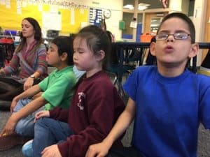 Un groupe d'enfants pratiquant la méditation en position assise, les yeux fermés et les mains posées sur les genoux.